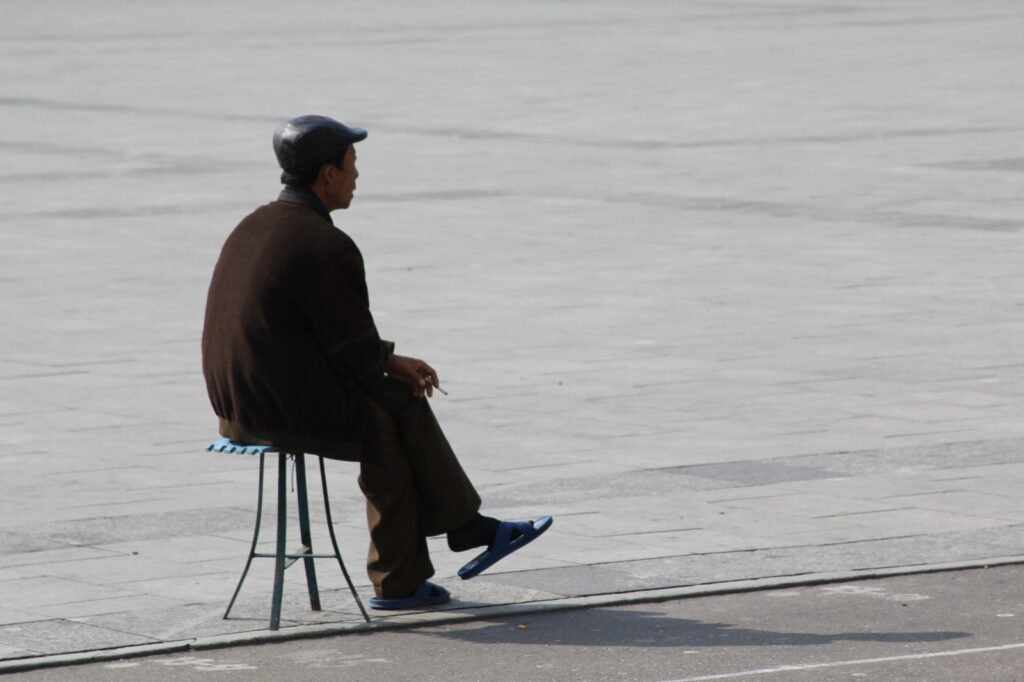 A man in slippers smoking on a stool in Kim Il-sung Square, Pyongyang, North Korea.